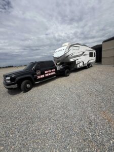 Home 26 MG branded truck towing white Cougar RV trailer on gravel, demonstrating RV hauling service